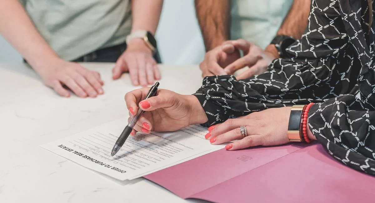 Close-up of hands signing a real estate document at a meeting table with three people.