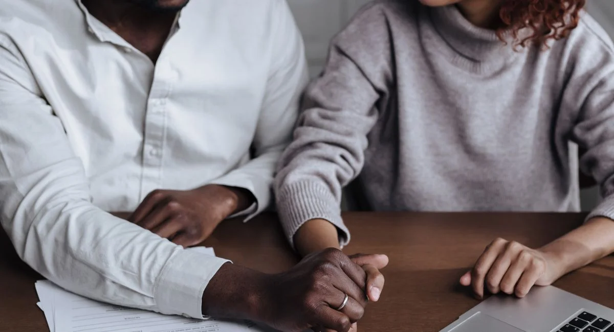 A diverse couple holding hands with a laptop and paperwork on the table.