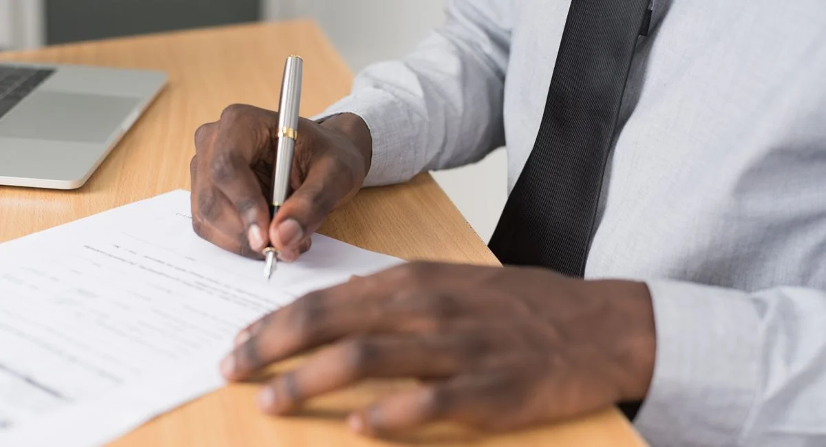 Close-up of a man signing paperwork on a desk, with a laptop nearby, in a professional office environment.