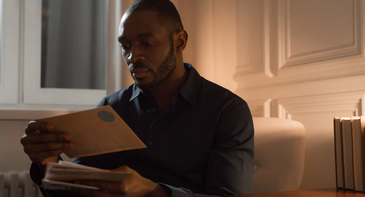 A man with a mustache reads letters while sitting in a warmly lit home interior.