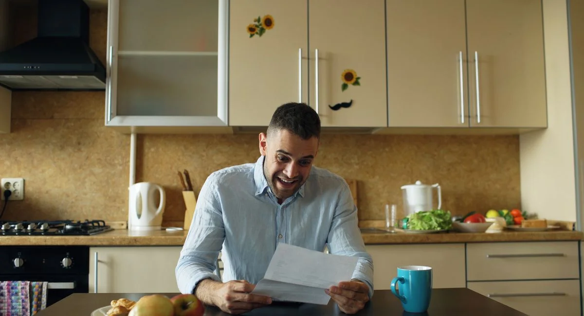 A delighted man reads a letter in a cozy kitchen setting, surrounded by food and drinks.