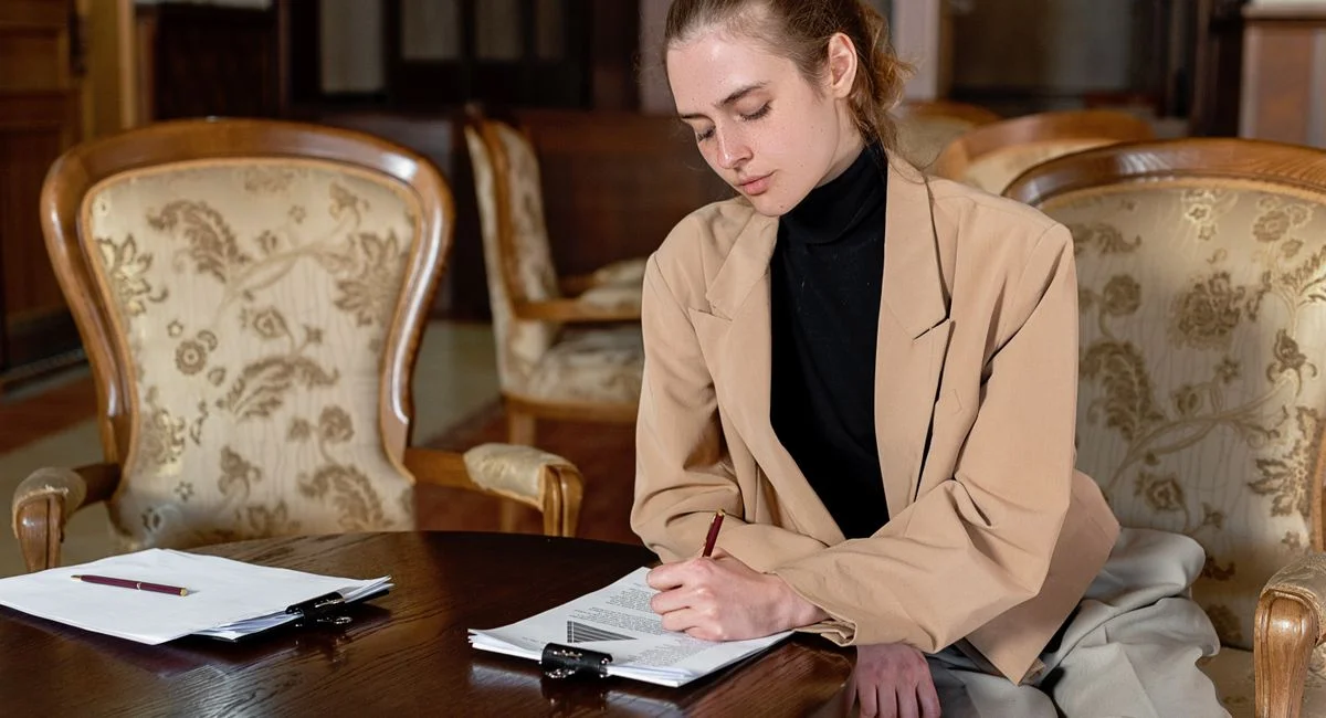 Young woman in a brown blazer signing documents at a table in a stylish office.
